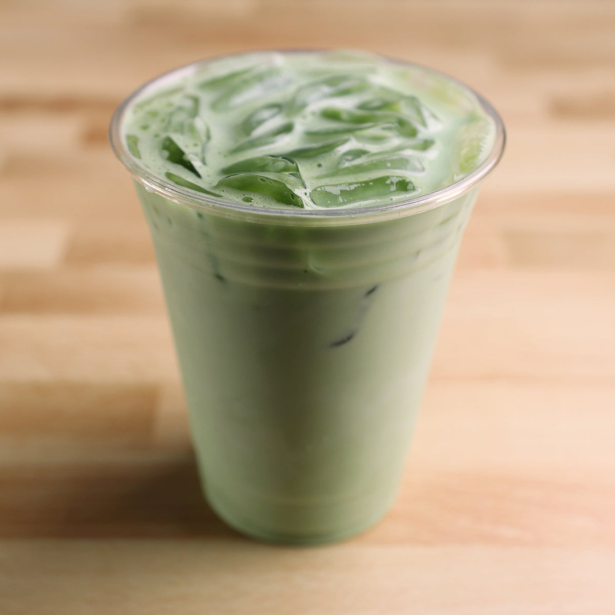 A clear plastic cup filled with vibrant green iced matcha, set on a light wood surface.