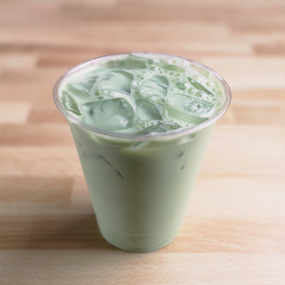 A clear plastic cup filled with vibrant green iced matcha, set on a light wood surface.